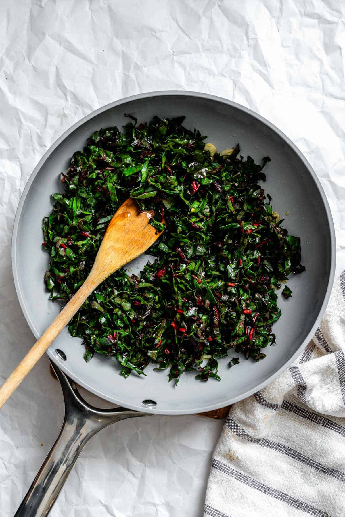 Overhead view of sautéed swiss chard in a gray skillet with wooden spoon, showing the greens wilted down with bits of red stems.