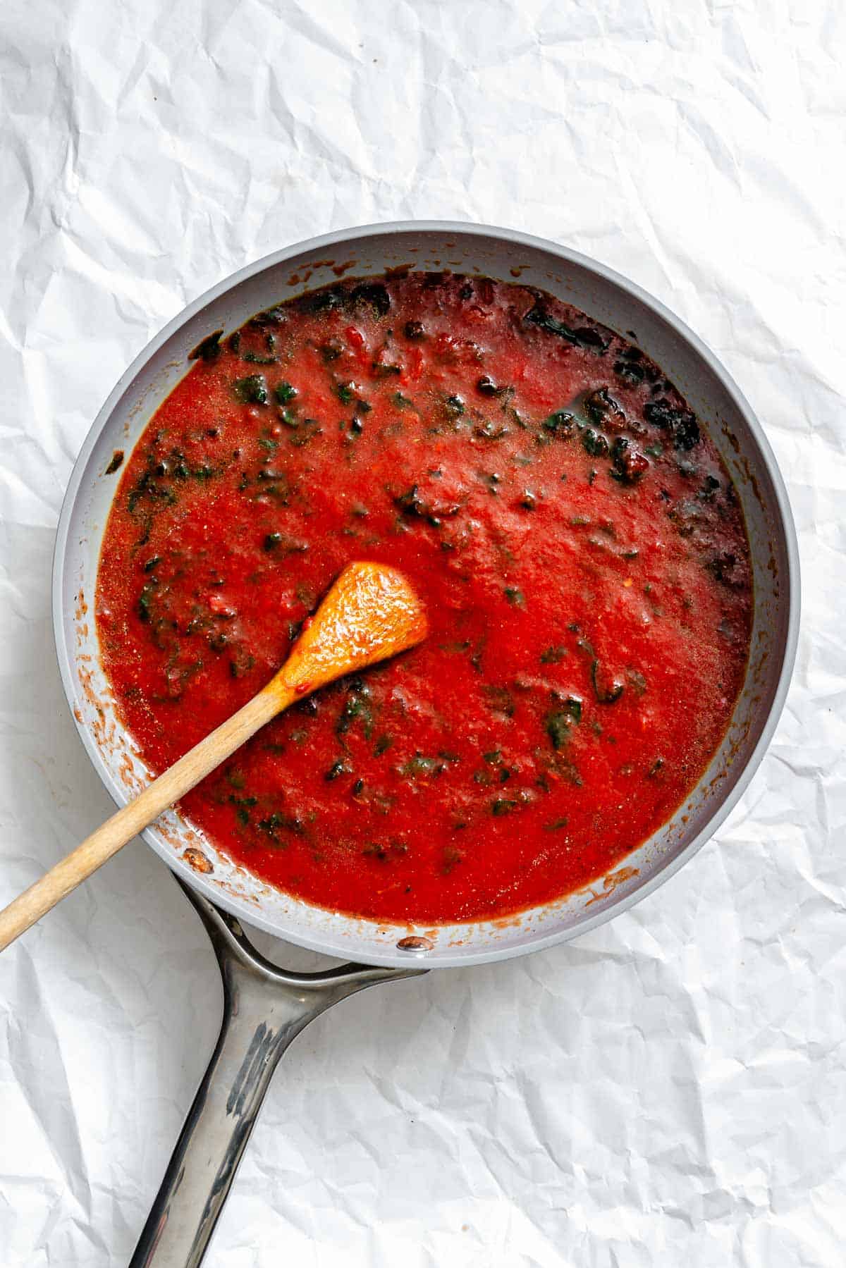 Overhead shot of tomato sauce simmering in a gray skillet with wooden spoon resting in the bright red sauce with herbs visible.