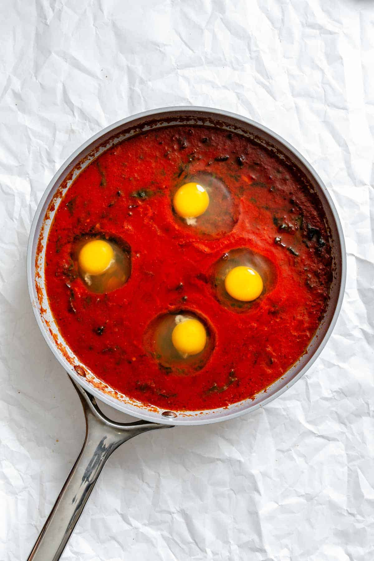 Overhead view of four raw eggs cracked into wells in the tomato sauce, creating indentations in the bright red mixture.