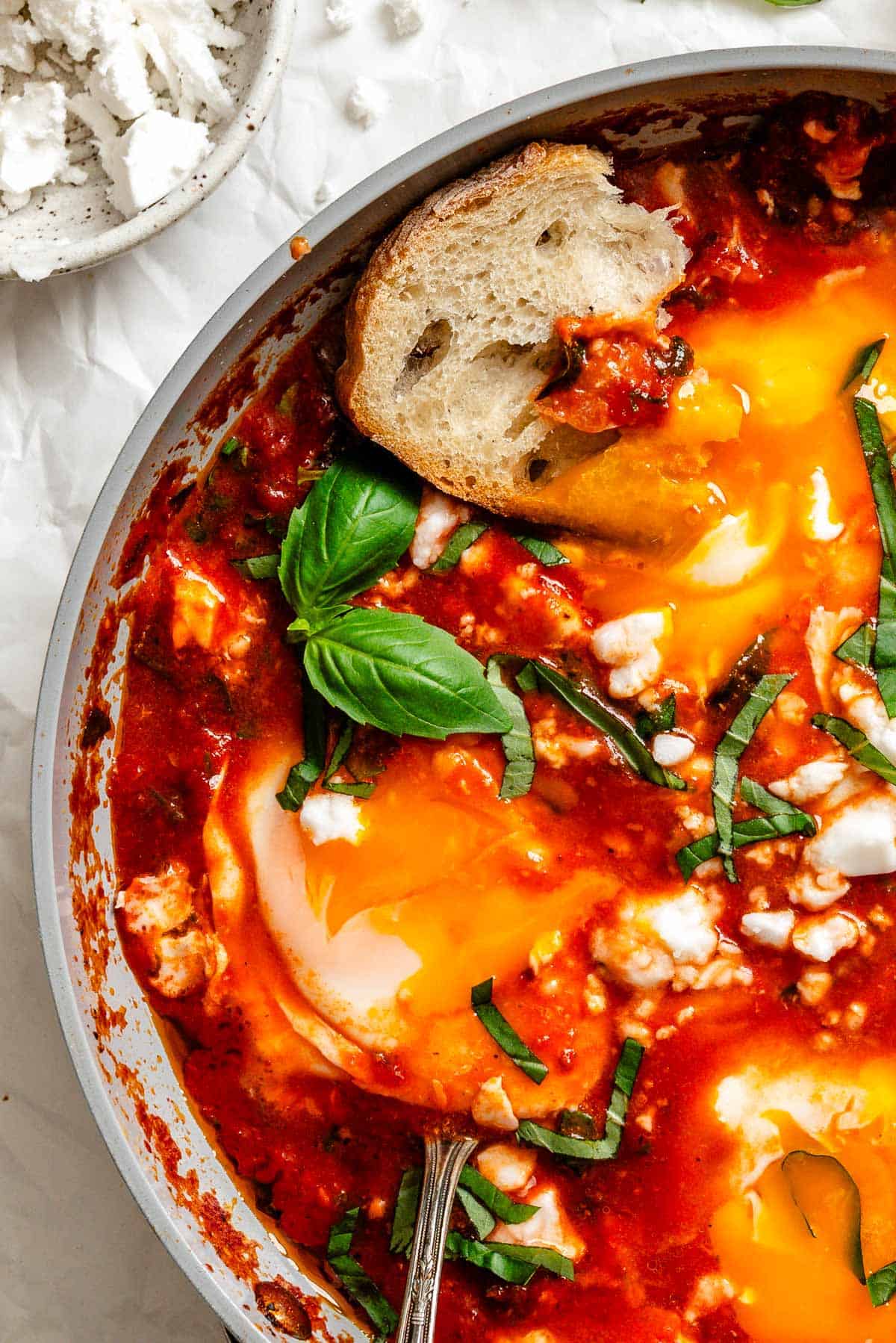Overhead view of bowl of crumbled goat cheese, chopped basil, and pan of eggs in purgatory with toasted bread on wooden board.