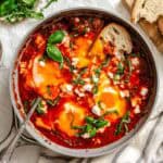 Finished eggs in purgatory in gray skillet with sliced rustic bread on wooden board, chopped basil and crumbled goat cheese in small bowls.