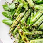 Overhead view of blistered green pea pods arranged on a white plate. The snap peas or English peas show caramelized edges from high-heat cooking with decorative pea tendrils and leaves as garnish. The bright green pods have a shiny, seasoned appearance.