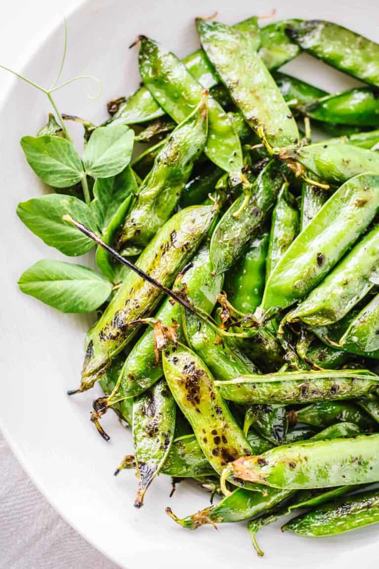 Overhead view of blistered green pea pods arranged on a white plate. The snap peas or English peas show caramelized edges from high-heat cooking with decorative pea tendrils and leaves as garnish. The bright green pods have a shiny, seasoned appearance.