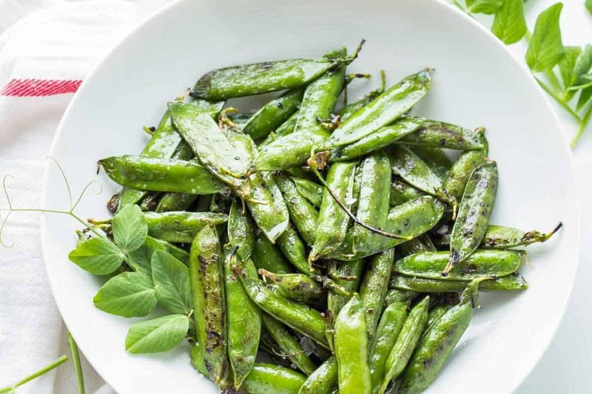 Close-up of roasted sugar snap peas or English peas in the pod on a white plate. The bright green pods show charred spots from roasting and are garnished with fresh pea shoots. The pods appear glossy, suggesting they've been tossed in oil.