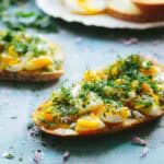 Several slices of toast topped with chopped soft-boiled eggs and fresh dill on a blue surface, with small purple flower petals scattered around.