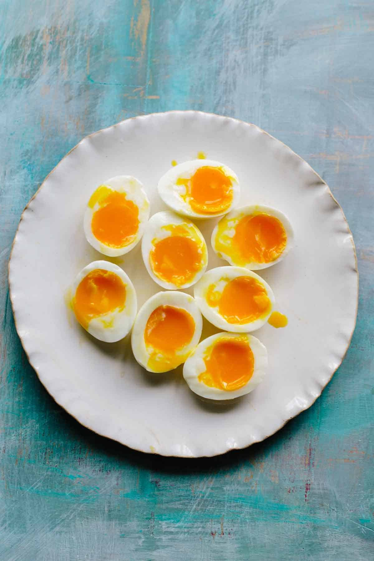 Perfect soft-boiled eggs cut in half showing bright orange-yellow jammy yolks arranged on a simple white plate against a turquoise blue surface.