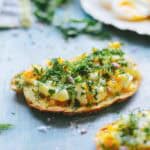 Close-up of toast topped with chopped soft-boiled eggs, fresh herbs, and seasonings on a blue surface with more eggs visible in the background.