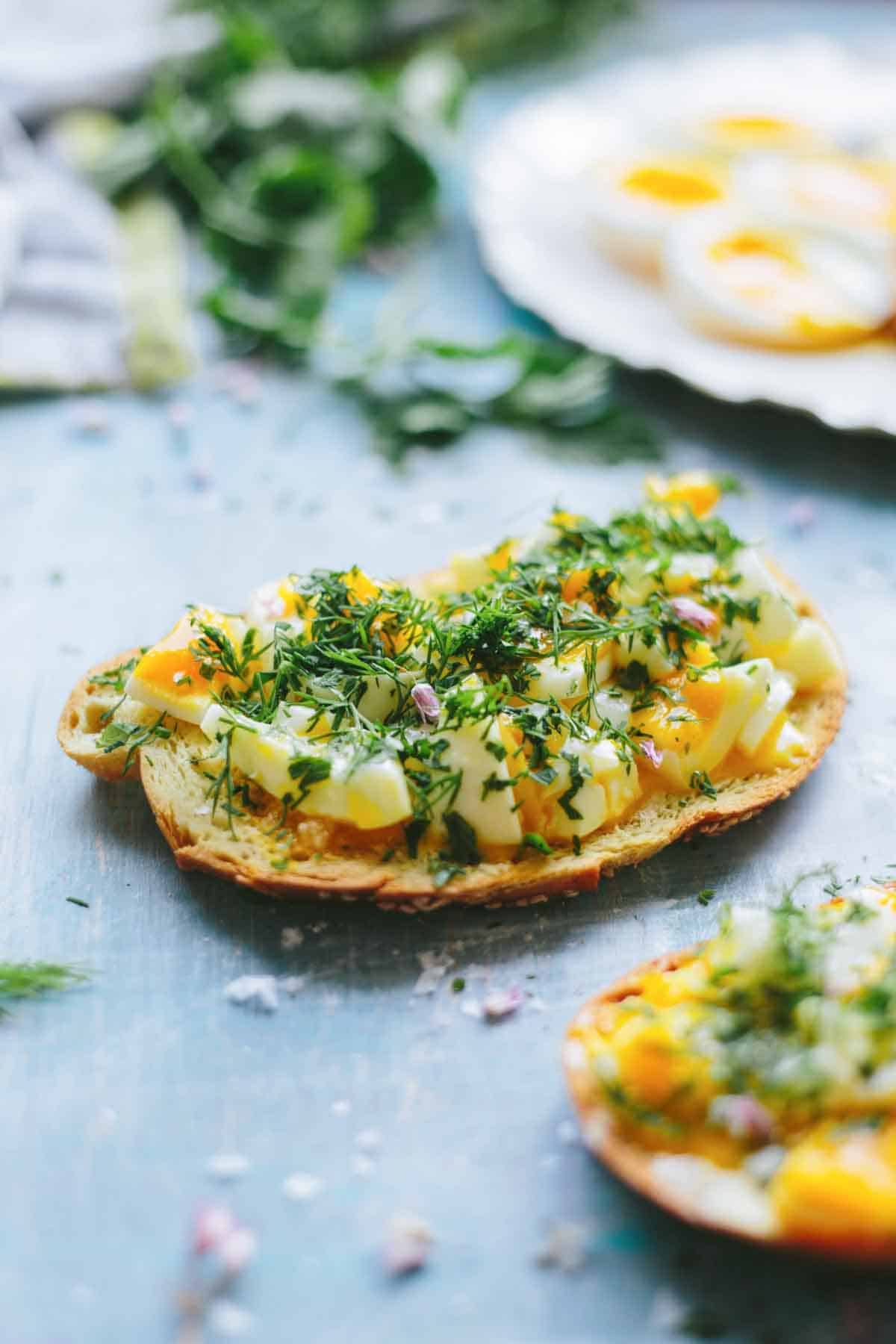 Close-up of toast topped with chopped soft-boiled eggs, fresh herbs, and seasonings on a blue surface with more eggs visible in the background.