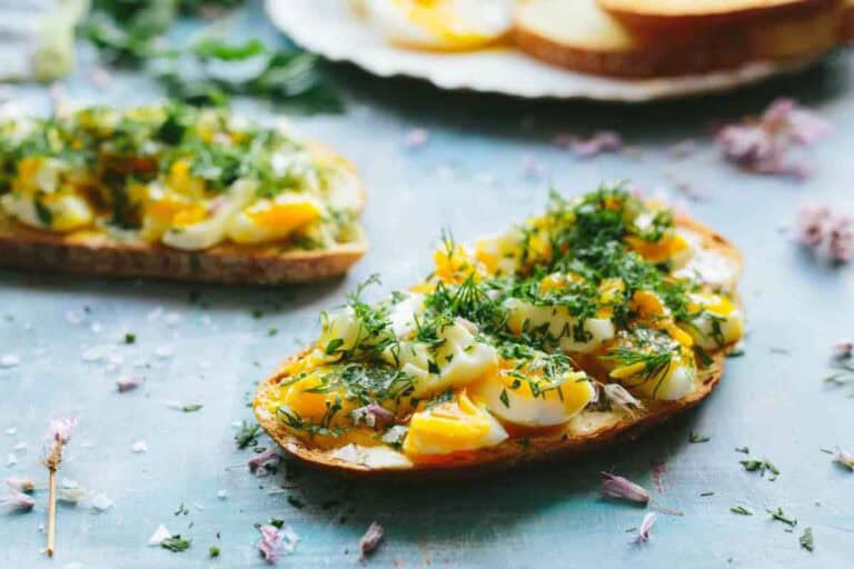Several slices of toast topped with chopped soft-boiled eggs and fresh dill on a blue surface, with small purple flower petals scattered around.