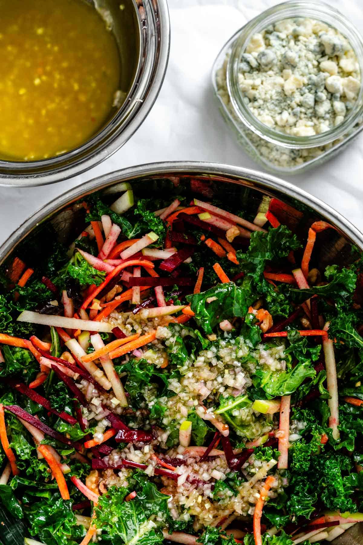 A close-up of a large mixing bowl containing combined salad ingredients - kale leaves mixed with colorful vegetable matchsticks and other elements, alongside a small jar of blue cheese and a bowl of dressing on a white background.