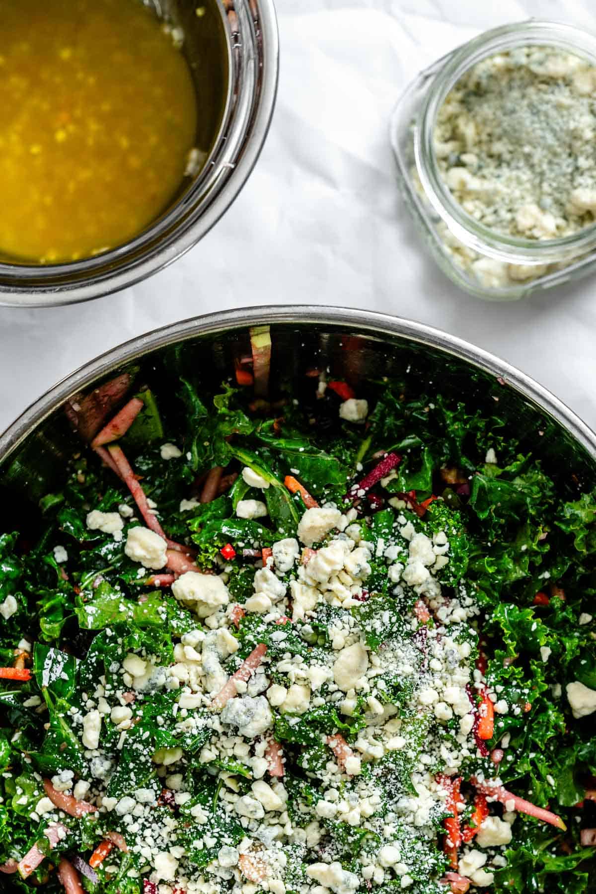 A similar angle of the mixing bowl now showing the fully dressed salad with crumbled blue cheese visible on top of the colorful vegetable and kale mixture.