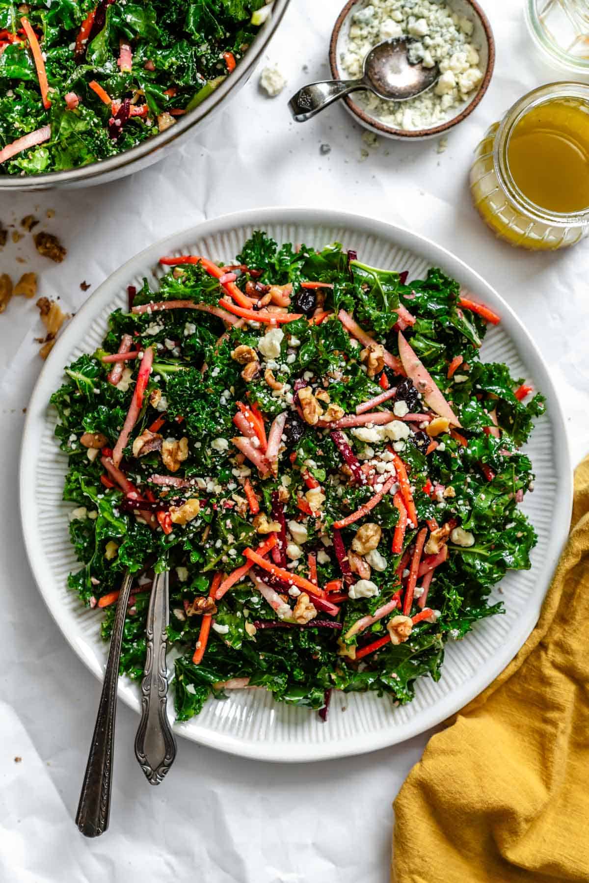 A white serving plate with the finished kale salad, beautifully arranged showing the vibrant colors of the vegetables, nuts, and cheese against the dark green kale backdrop. A small bowl of blue cheese and a jar of dressing are visible to the side.