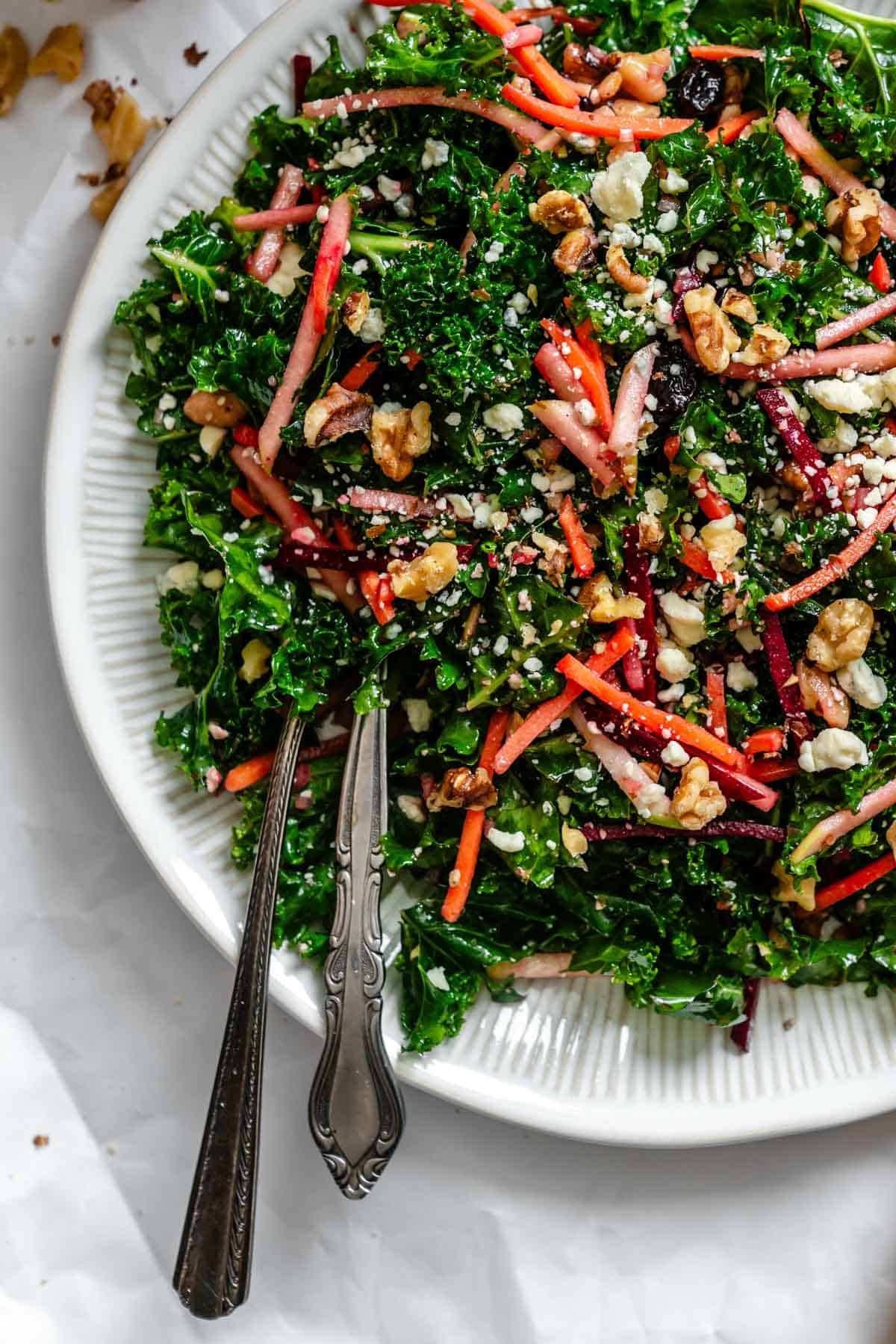 Another view of the plated kale salad with serving utensils, showing its texture and colorful ingredients, with blue cheese crumbles, walnut pieces, and julienned vegetables mixed throughout the kale leaves.