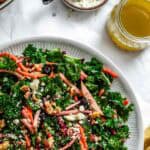An overhead view of a white plate with the fall harvest kale salad alongside another serving, with small containers of blue cheese and dressing visible, all arranged on a white surface with a mustard yellow napkin.