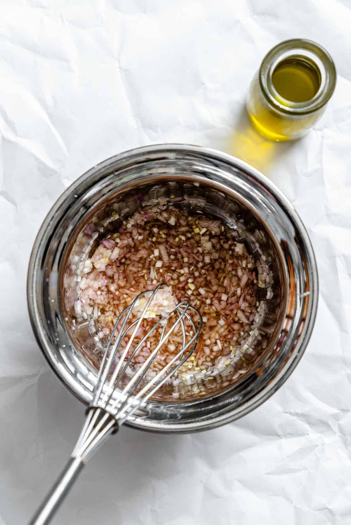 A stainless steel mixing bowl containing a vinaigrette in progress with finely minced shallots soaking in liquid with a metal whisk, all set on white parchment paper with a small bottle of olive oil visible in the corner.
