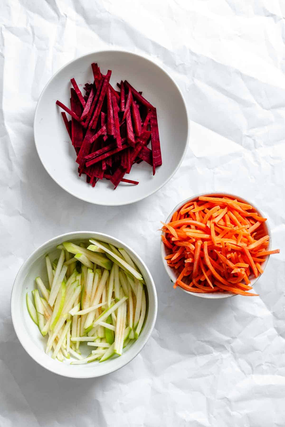 Three white bowls containing julienned vegetables on white parchment paper - one with bright red beet matchsticks, one with pale green apple matchsticks, and one with orange carrot matchsticks, creating a vibrant color palette.
