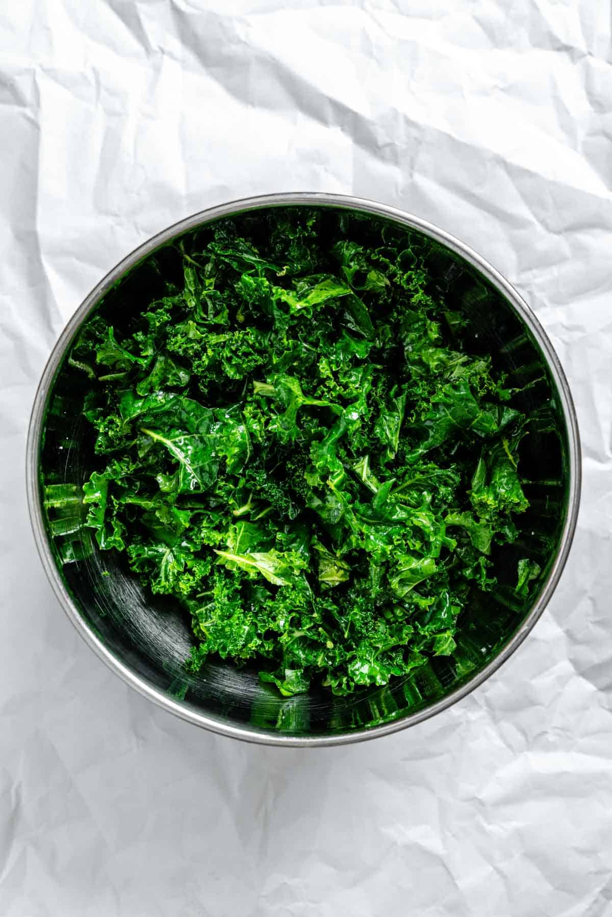 A stainless steel bowl filled with fresh, dark green kale leaves on white parchment paper. The leaves appear to be washed and ready for preparation in a salad.