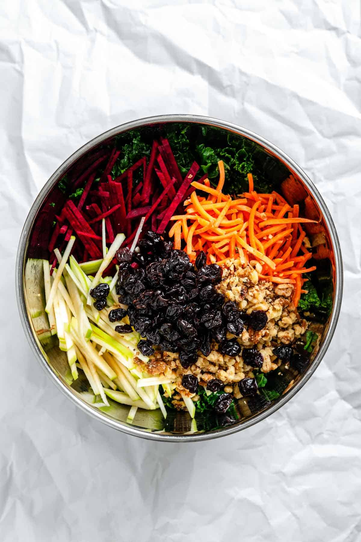 A bird's eye view of a stainless steel mixing bowl containing julienned vegetables (red beets, green apples, orange carrots) arranged in sections atop kale leaves, with a pile of dried cranberries and chopped walnuts in the center.