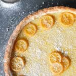 Closeup of a golden Meyer lemon tart with thin lemon slices arranged on a creamy yellow filling, dusted with powdered sugar. A small mesh sifter with powdered sugar is visible in the corner on a dark countertop.