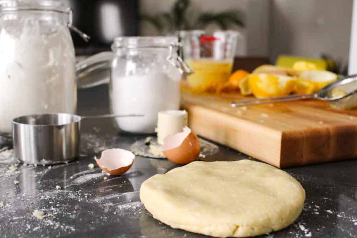 Baking ingredients for lemon tart preparation including glass jars of flour and sugar, a disc of pie dough, butter, egg shells, and sliced lemons on a wooden cutting board on a dark surface.