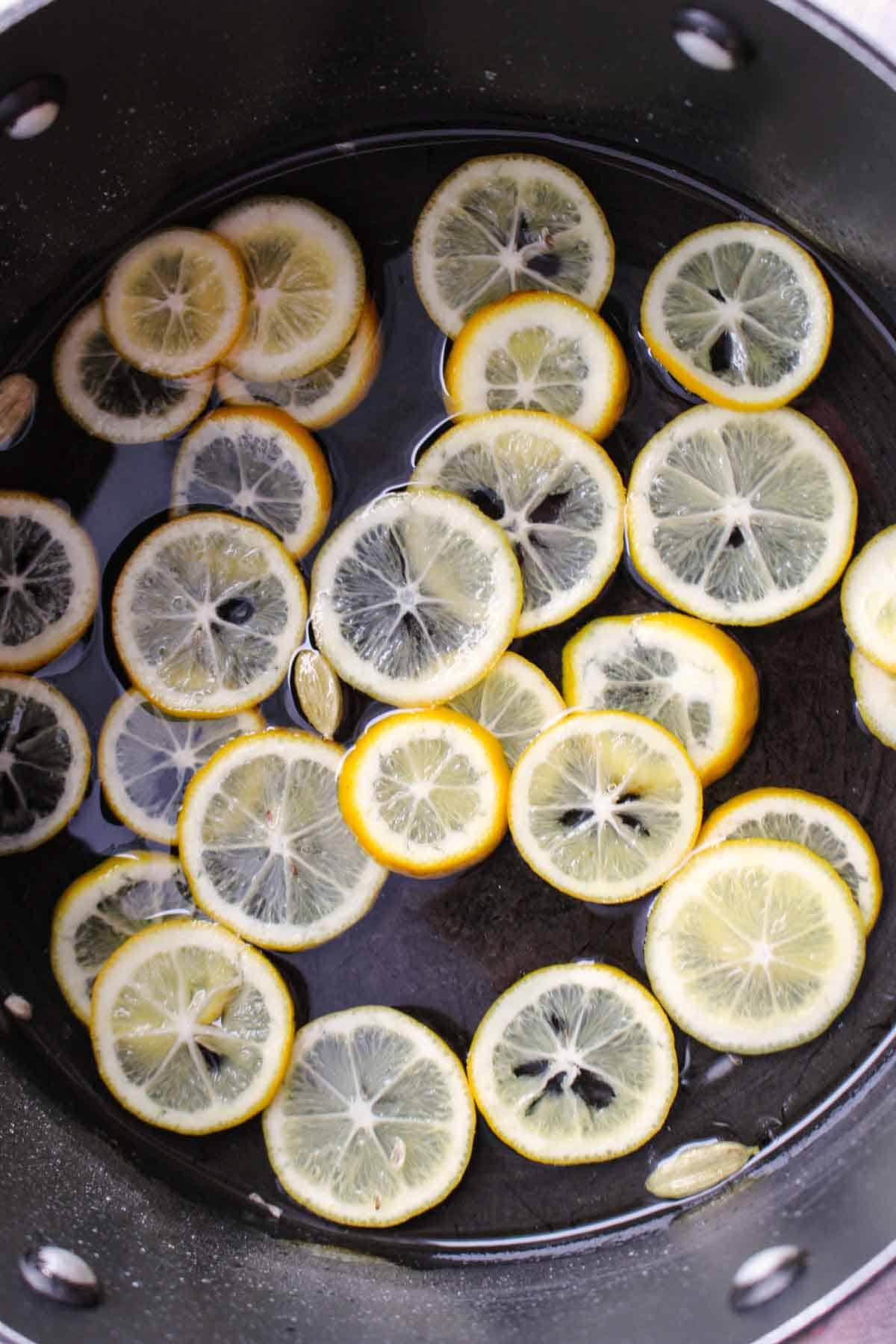Thinly sliced Meyer lemons floating in syrup in a black saucepan, showing the translucent lemon wheels with their star-like centers during the candying process.