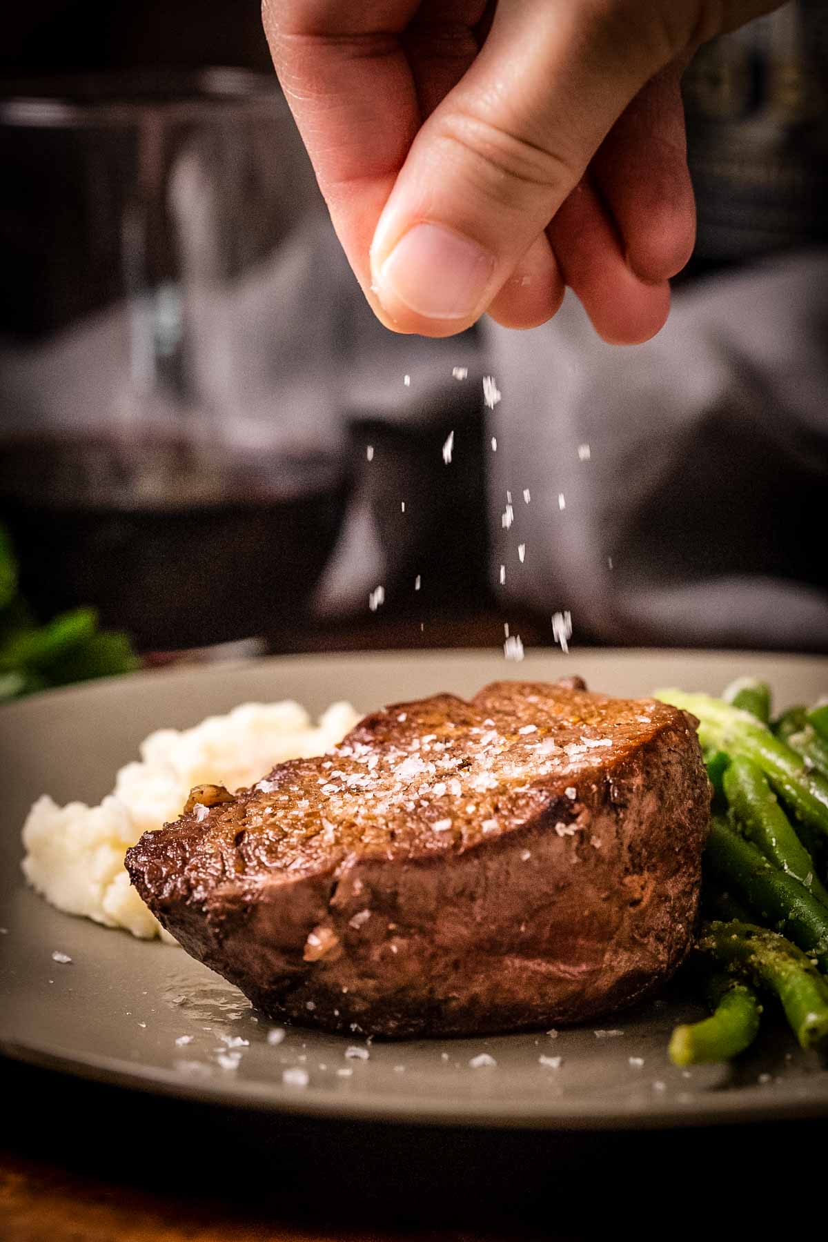 A hand sprinkling salt over a cooked filet mignon plated with mashed potatoes and green beans. The steak is the focal point on a gray plate with the seasoning caught in mid-air.