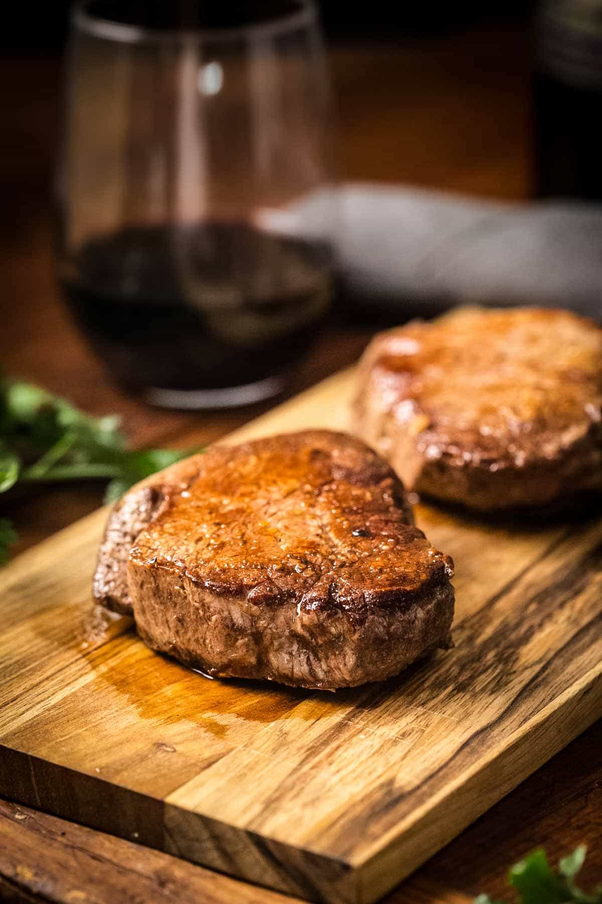 A close-up of two filet mignon steaks on a wooden serving board. The steaks are photographed from an angle that highlights their caramelized crust and juicy interior, with a glass of red wine blurred in the background.