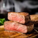A dramatic close-up of two stacked filet mignon steaks with one cut open to display its perfectly medium-rare center. The photo captures the contrast between the seared exterior and red interior.
