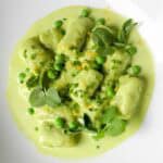 Pillowy green gnocchi served in a creamy light green sauce with fresh peas, microgreens, and orange zest on a white plate.