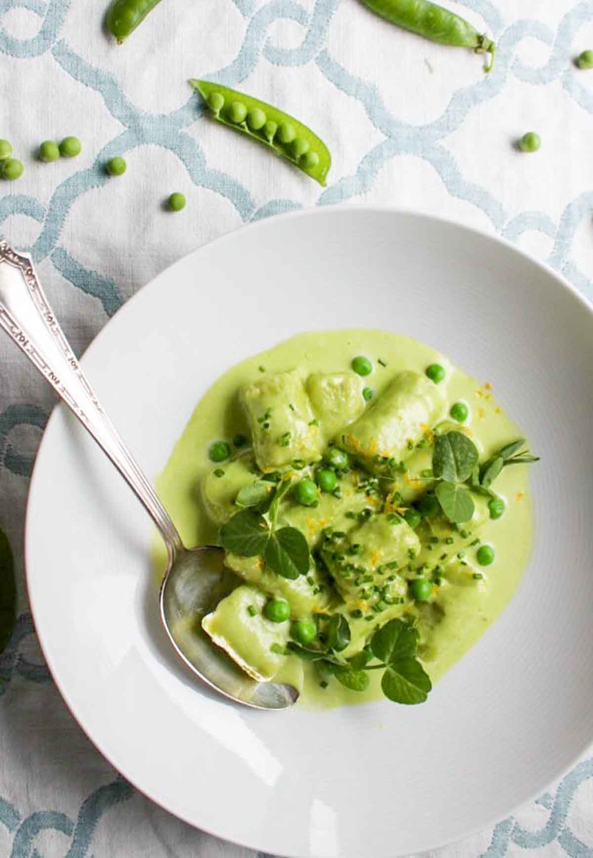 Overhead view of spring pea gnocchi in a white bowl with fresh peas and pea shoots, with whole pea pods visible in the background.