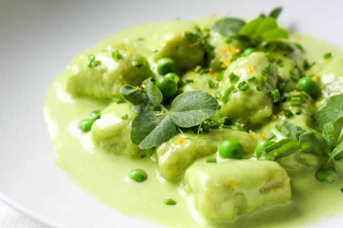 Close-up detail of spring pea gnocchi with their characteristic ridges in a pale green sauce garnished with fresh pea shoots and herbs.
