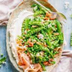 Overhead view of an elegant oval serving platter with bright green peas and snap peas topped with prosciutto and fresh herbs, served with a swirl of creamy ricotta cheese.