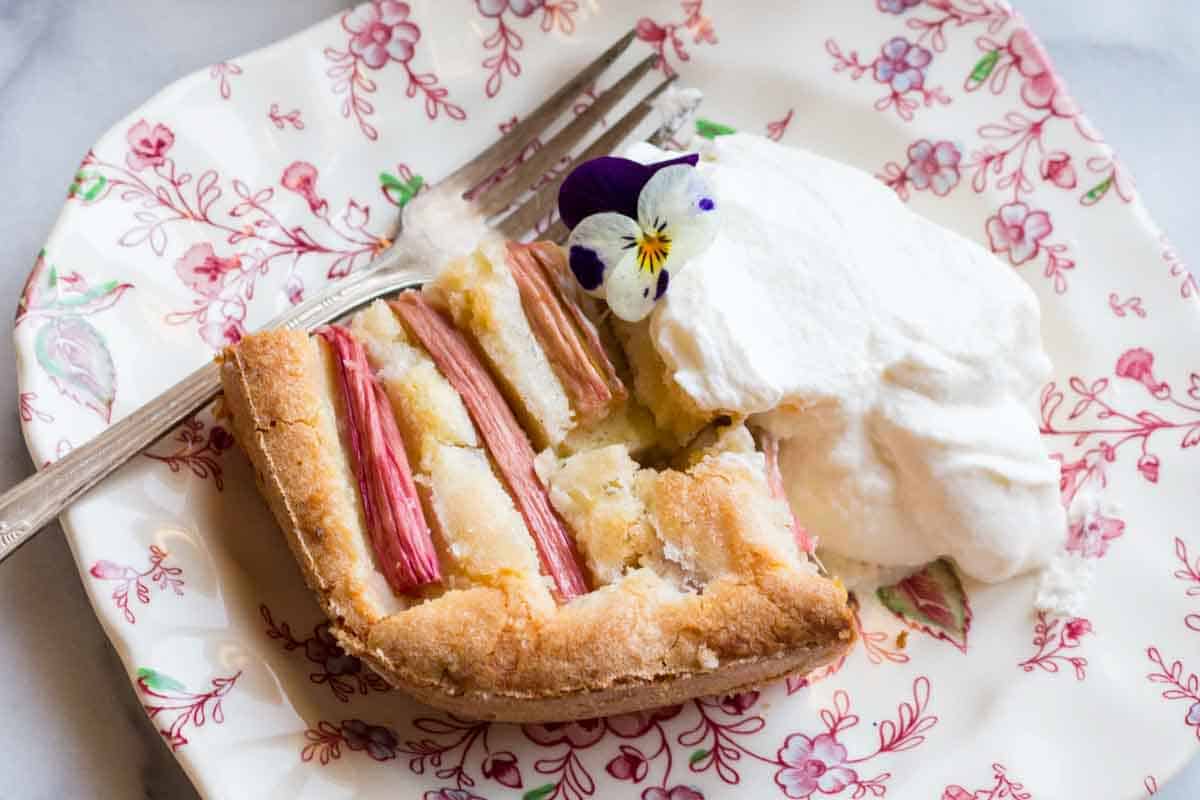 A slice of rhubarb cake served on a decorative floral plate with a dollop of whipped cream and an edible flower garnish.