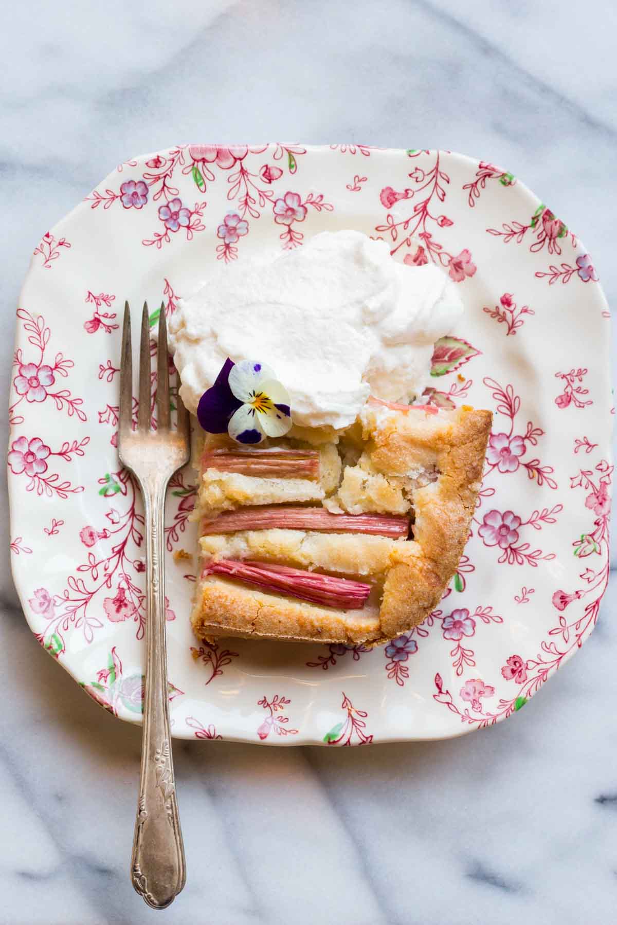 Overhead view of a slice of rhubarb cake on a floral plate with whipped cream and an edible purple pansy flower.