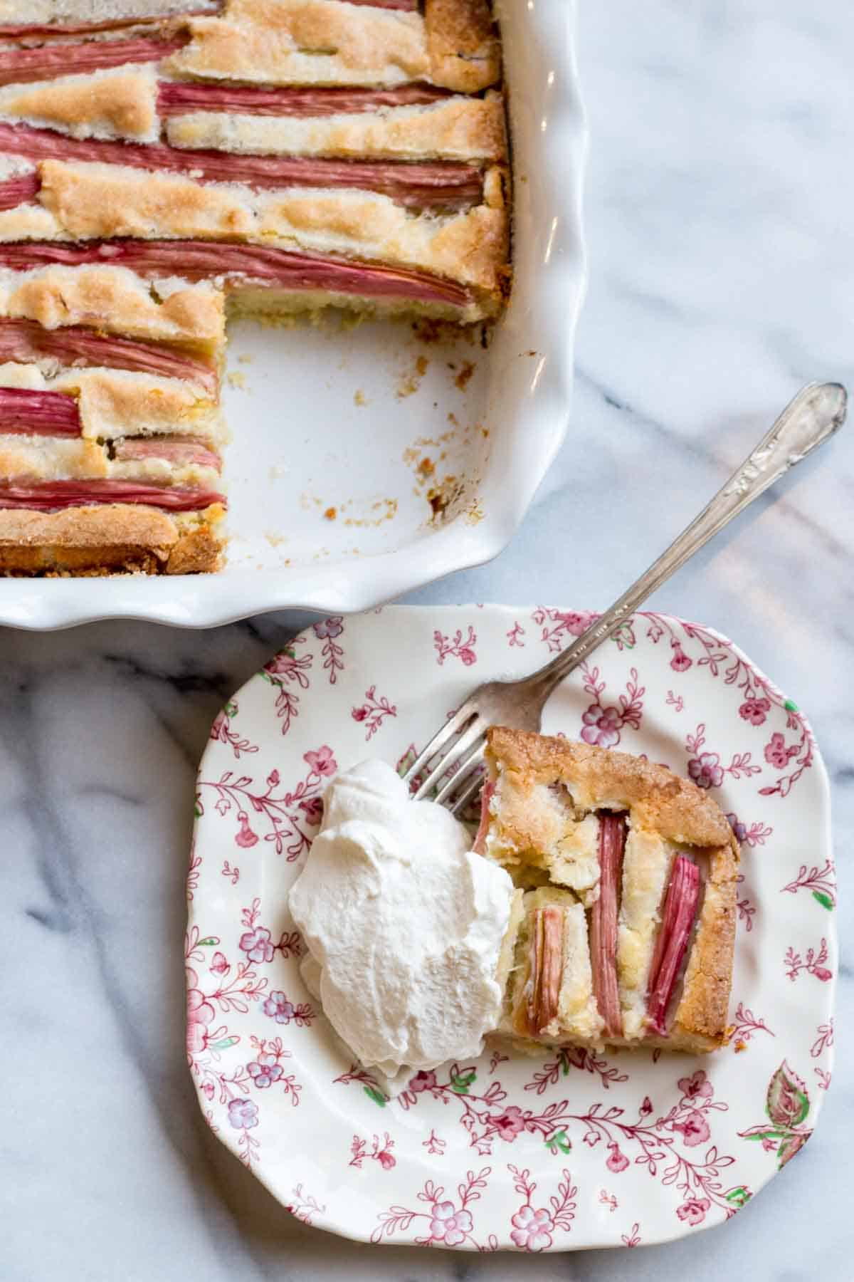 Top-down view of a rhubarb cake in a white ceramic dish with a slice removed, showing the contrast between the cake and pink rhubarb stalks.