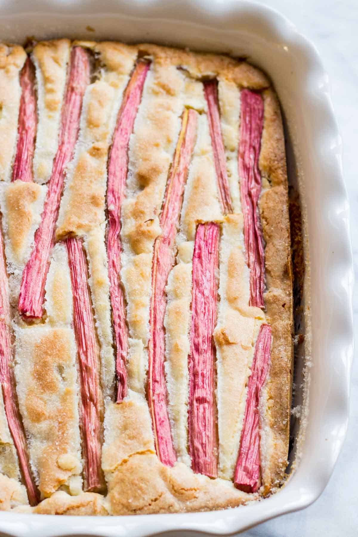 Close-up view of a rhubarb cake in a baking dish showing bright pink rhubarb stalks embedded in golden cake batter.
