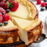 Close-up view of a ricotta cheesecake with a slice being removed, showing the smooth, dense texture. The cake is decorated with fresh berries and the golden brown crust is visible along the sides.
