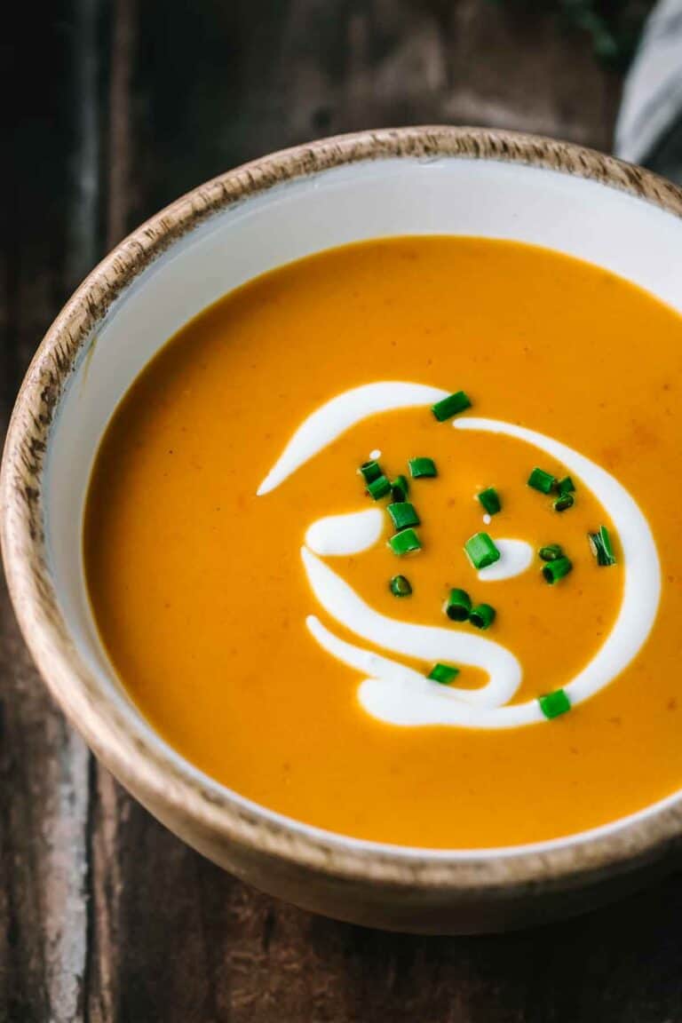 Extreme close-up of shrimp bisque in a wooden-rimmed white bowl showing the smooth, velvety orange-colored soup with decorative cream swirl and bright green chive garnish against a dark wooden background.