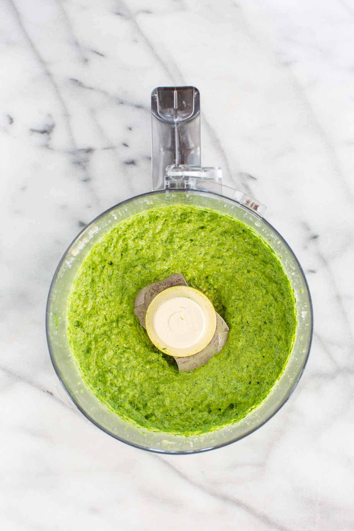 Bright green spinach pesto freshly blended in a food processor bowl on a white marble surface, showing smooth, vibrant texture.
