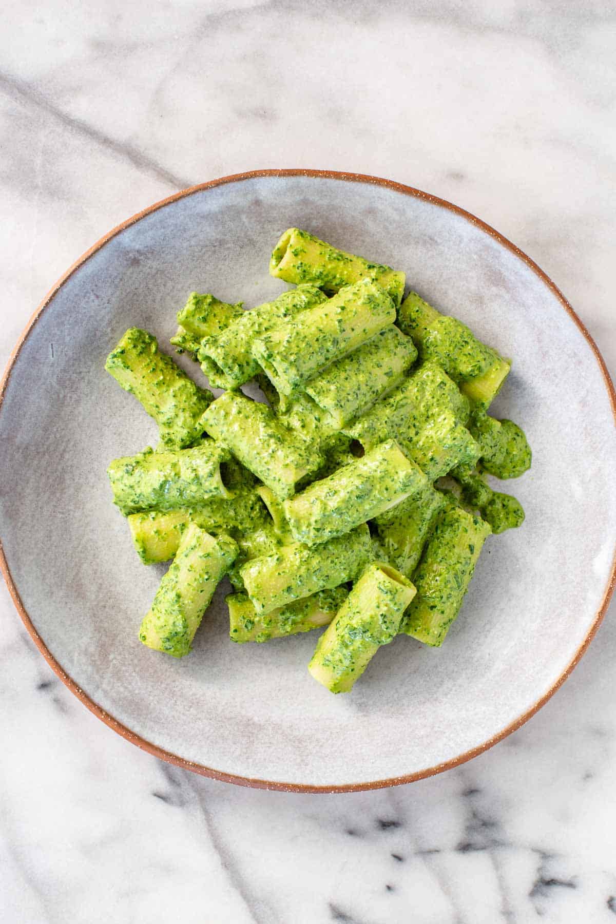 Overhead view of rigatoni pasta coated in vibrant green spinach pesto, served on a light gray ceramic plate against a white marble background.