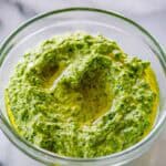 Glass bowl of finished spinach pesto with a pool of olive oil on top, showing the rich, vibrant green color and creamy texture.