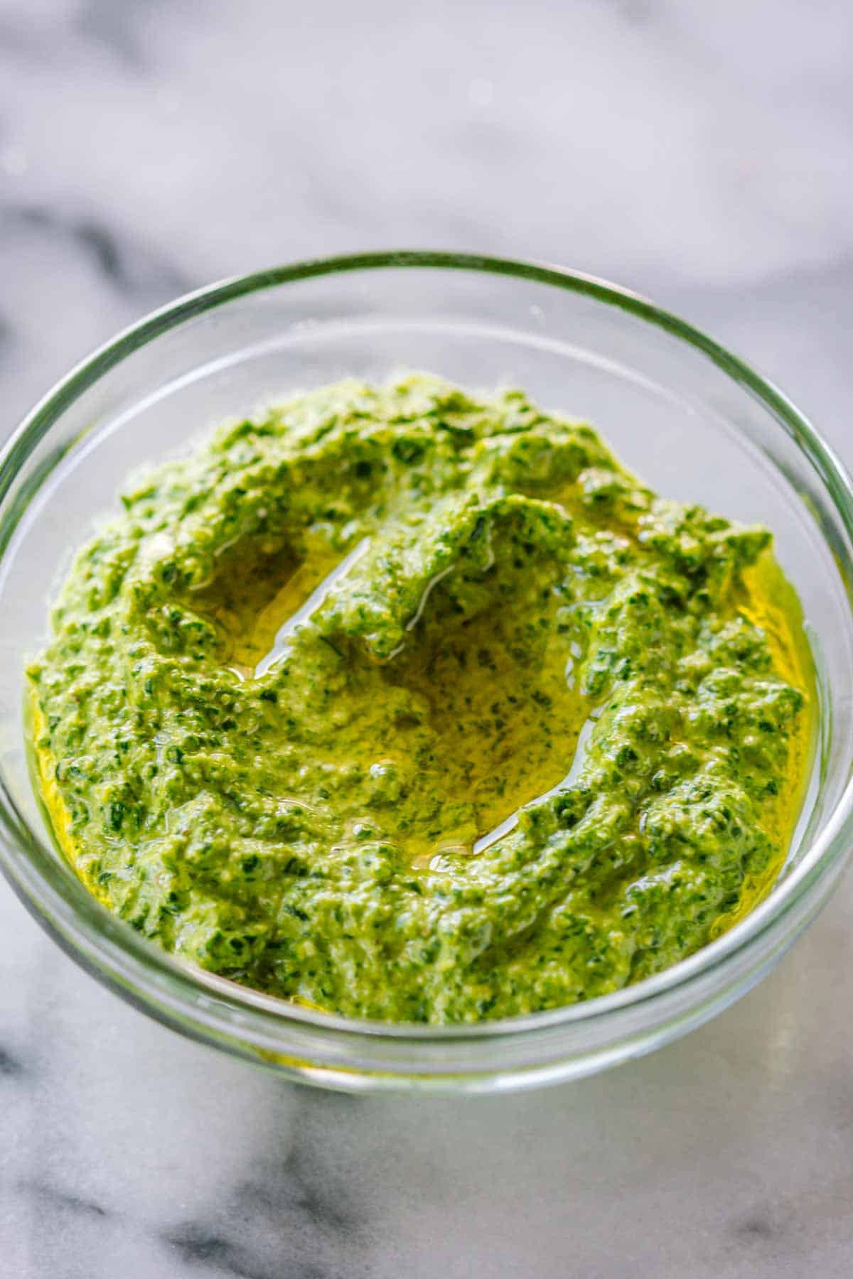 Glass bowl of finished spinach pesto with a pool of olive oil on top, showing the rich, vibrant green color and creamy texture.