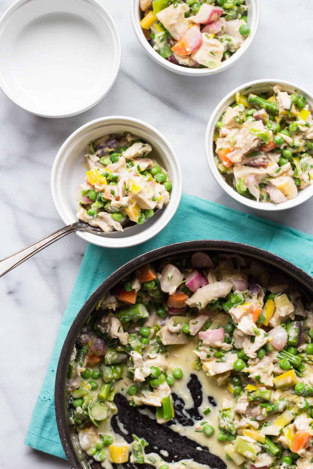 Overhead view of a creamy chicken pot pie filling in a black skillet with three individual servings in white ramekins. The filling contains chicken, peas, carrots, and colorful vegetables in a light cream sauce. A spoon shows the texture of the filling in one ramekin.