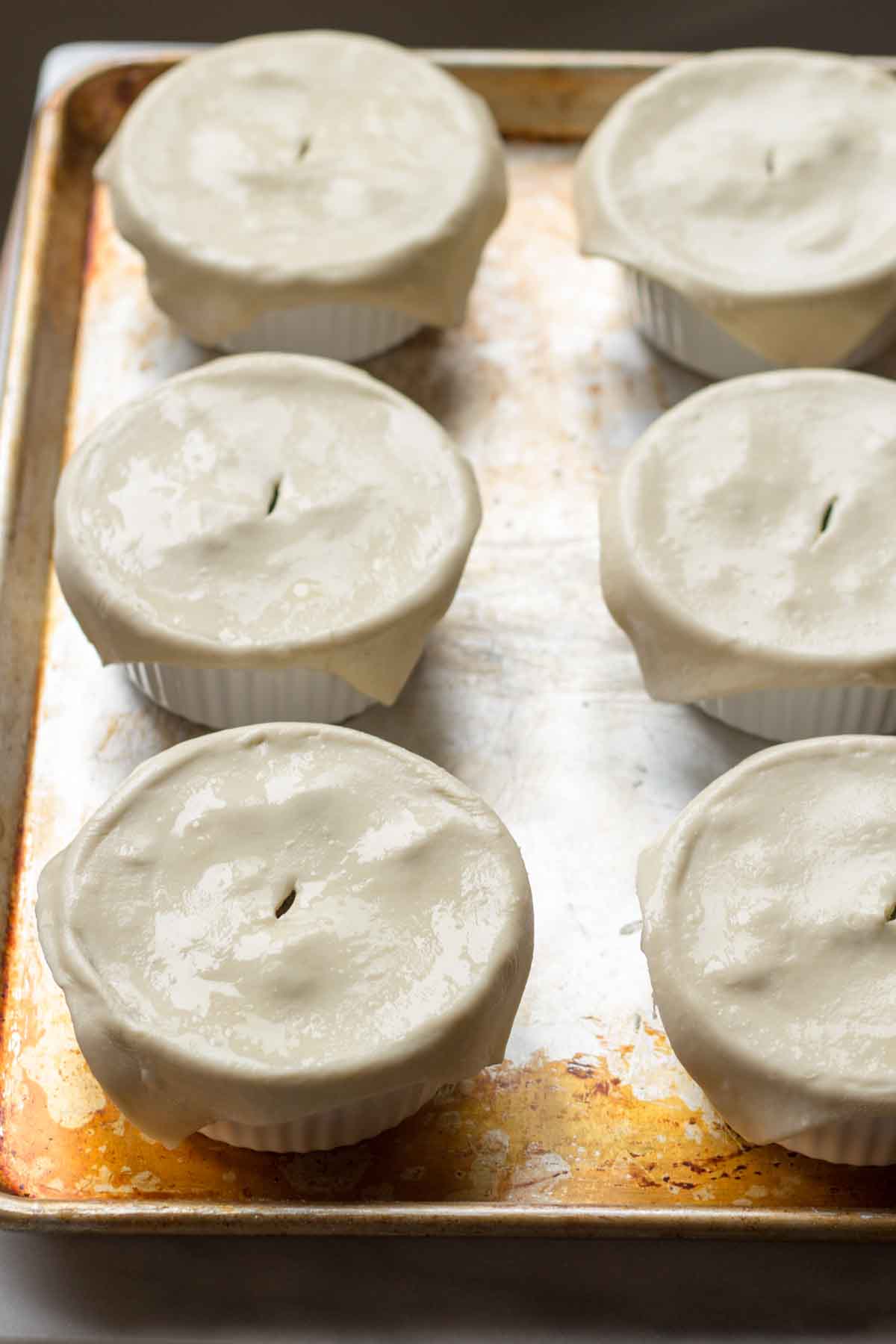 Unbaked individual chicken pot pies in six white ramekins on a baking sheet. Each ramekin is covered with raw pastry dough that has a small vent hole in the center, ready to be baked.