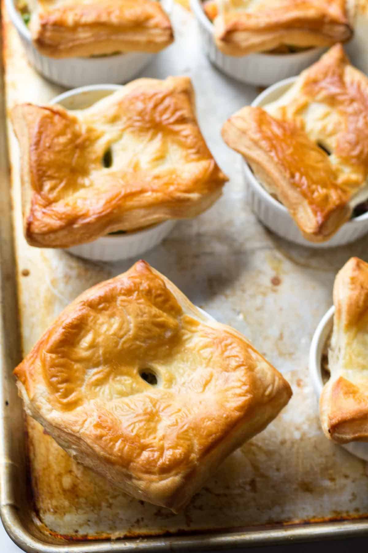 Freshly baked individual chicken pot pies in white ramekins on a baking sheet. The puff pastry tops are golden brown and flaky with small vent holes in the center.
