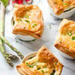 Four golden-brown chicken pot pies in white ramekins on a marble surface, garnished with fresh chives. Fresh asparagus spears and radishes are visible as decorative elements. The puff pastry tops are beautifully puffed and flaky.