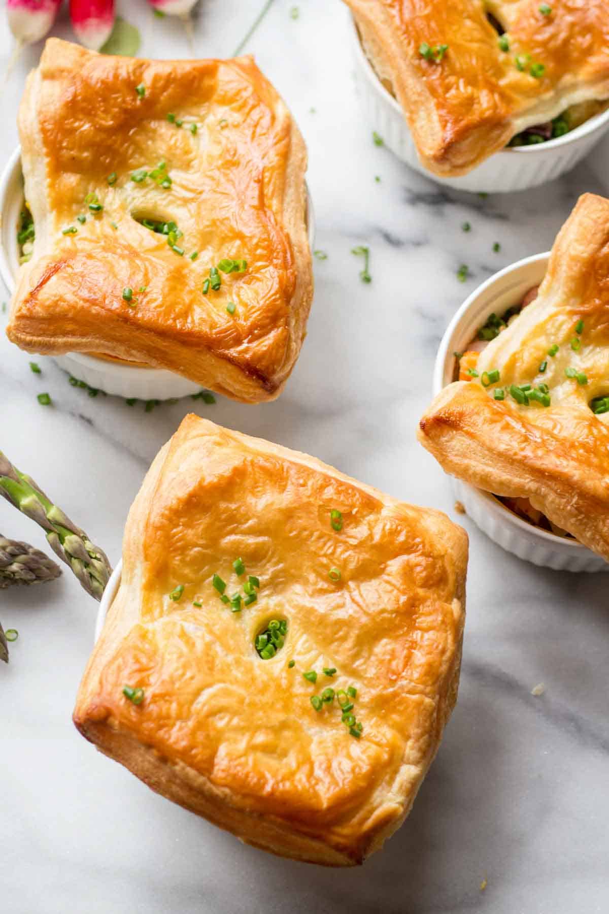 Close-up of golden-brown chicken pot pies in white ramekins on a marble surface. The pastry tops are decorated with minced chives, and the flaky, buttery crust shows beautiful layering.