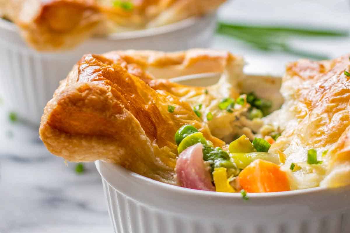 Close-up of an individual chicken pot pie with the golden puff pastry top broken open, revealing the creamy filling of chicken, peas, carrots and vegetables inside. The pastry is flaky and golden brown.