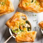 Overhead view of an individual chicken pot pie with part of the golden pastry top broken and a spoonful of filling being lifted out. The filling shows pieces of chicken, colorful vegetables, and a creamy sauce.