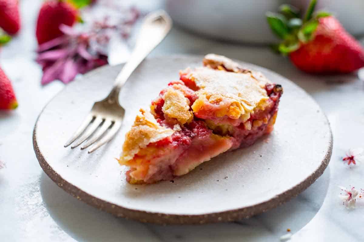 A slice of strawberry pie on a white ceramic plate with a fork beside it. The pie has a golden crust and vibrant red strawberry filling. Fresh strawberries and purple flowers are visible in the background.